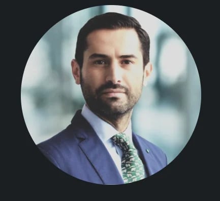 Professional headshot of a bearded man in a blue suit and patterned tie against a blurred background